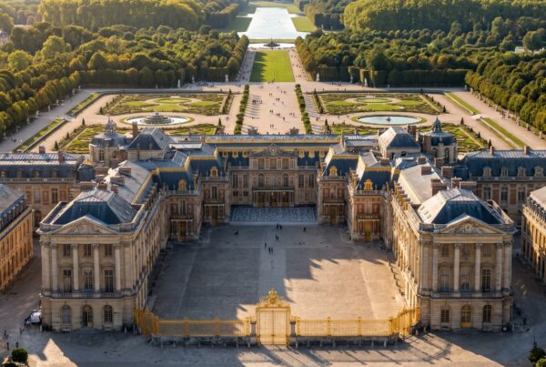 Aerial view of the Palace of Versailles featuring the golden Royal Gates, Hall of Mirrors wing, formal French gardens, fountains, and the Grand Canal on a clear sunny day